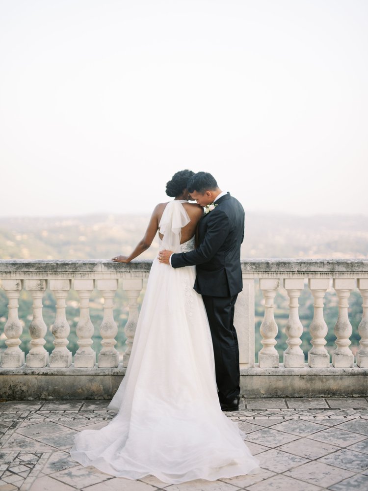 A bride and groom stand on a stone balcony overlooking a hazy landscape. The bride, wearing a white gown, leans on the railing while the groom, in a dark suit, embraces her. They face away from the camera, sharing an intimate, serene moment.