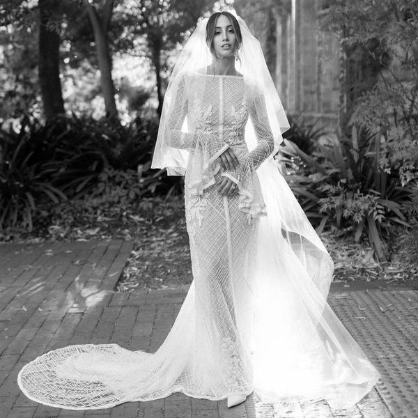 A bride stands outdoors in a lush garden setting, wearing a detailed, long-sleeve lace wedding dress with a long train and veil. Sunlight filters through trees, creating a serene and ethereal atmosphere. The black-and-white photo captures the elegance of the scene.