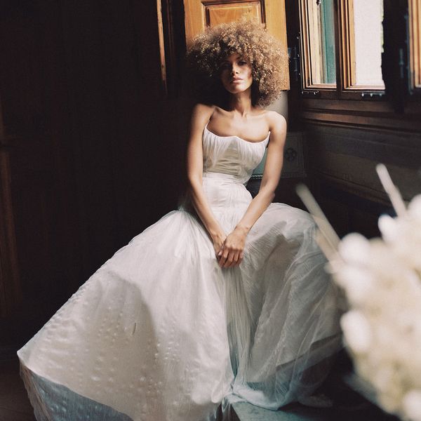 A woman with curly hair sits by a window in a dimly lit room, wearing a white, strapless wedding gown with a voluminous skirt. She has a contemplative expression and is surrounded by wooden paneling and soft natural light.