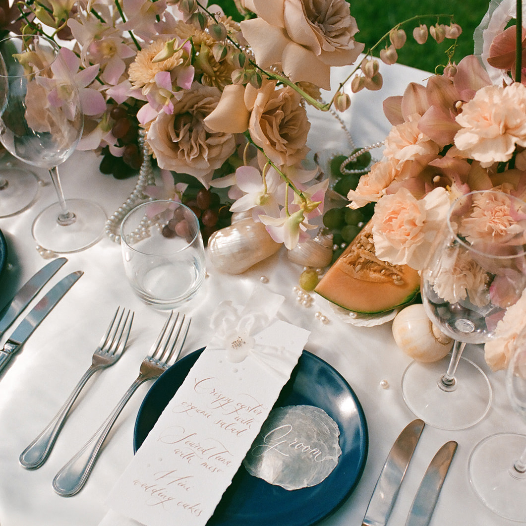 A beautifully set dining table with elegant pink and peach floral arrangements, candles, and fruits. Each place setting includes a blue plate, silver flatware, wine glasses, and a handwritten menu card with delicate calligraphy resting on a white napkin.