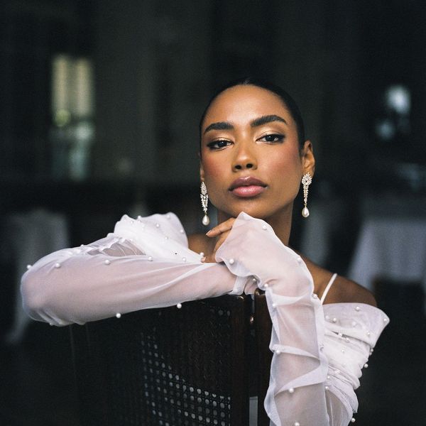 A young woman with a serene expression poses indoors. She is resting her chin on her hand while leaning against a chair. She wears a white, pearl-embellished, sheer sleeved dress and elegant pearl drop earrings. The background is softly lit and blurred.