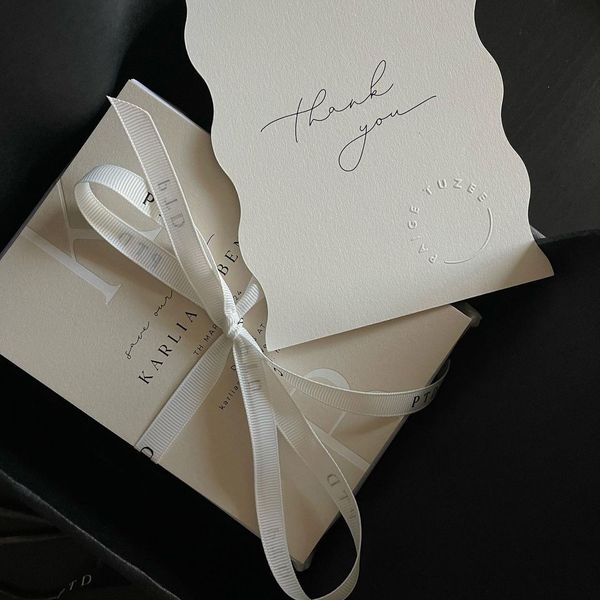 A white box tied with a white ribbon sits in black surroundings. On top of the box is a white card with a scalloped edge, featuring the handwritten words "thank you." The card features the logo "FRAGRANCE CAFE" embossed on it.