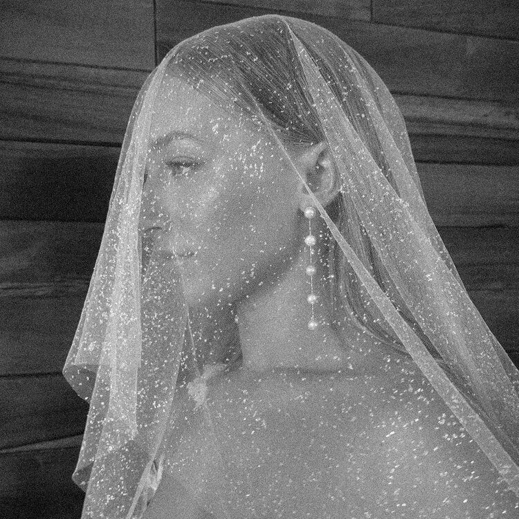 Black and white photo of a bride with a sparkling veil covering her face. She wears long, dangling pearl earrings and has her hair pulled back. The background is comprised of wooden planks. The overall ambiance is elegant and serene.