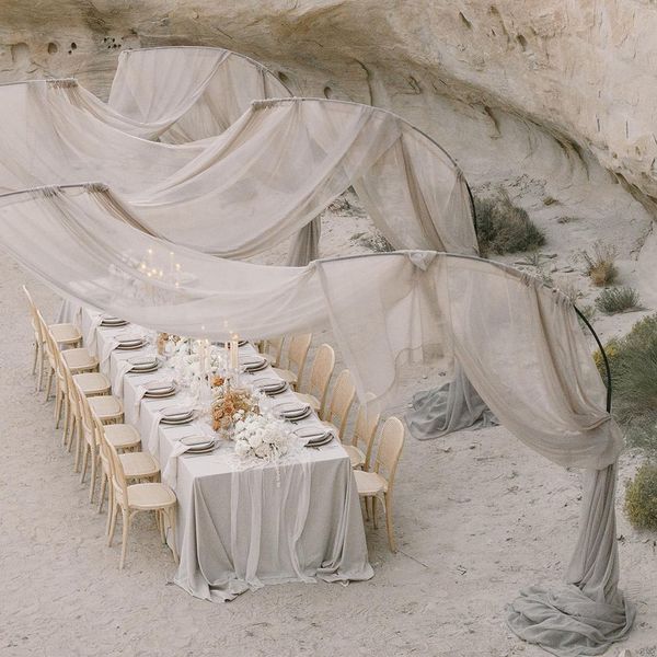 A long, elegant dining table is set in a rocky, desert-like environment. The table is covered with a light grey cloth and adorned with white floral centerpieces and candles. Sheer, billowing fabric drapes from the rocky walls above, creating an ethereal canopy.