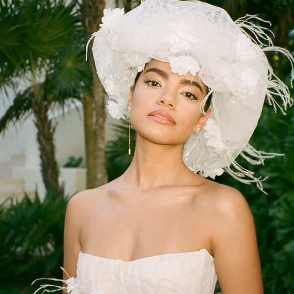 A woman wearing an elegant white off-shoulder dress and a large white floral hat poses outdoors. The background features tropical greenery and palm trees. Her expression is calm and confident.