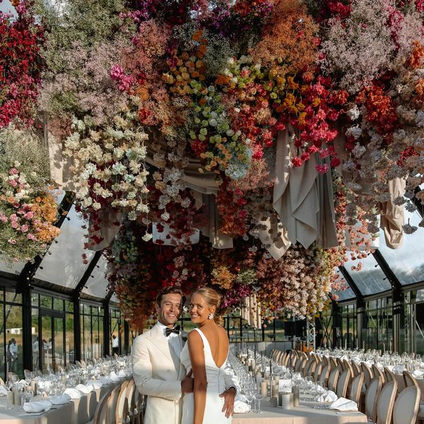 A couple in elegant white attire stands beneath a stunning floral ceiling installation. The flowers are a vibrant mix of colors, creating a lush canopy over the long dining tables arranged for an event in a spacious, glass-roofed venue.