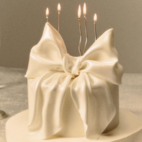 A small, elegant cake adorned with smooth white fondant that is sculpted to resemble a large, intricate bow. Five lit candles are placed on top, their flames gently flickering, adding a warm, celebratory glow to the scene.