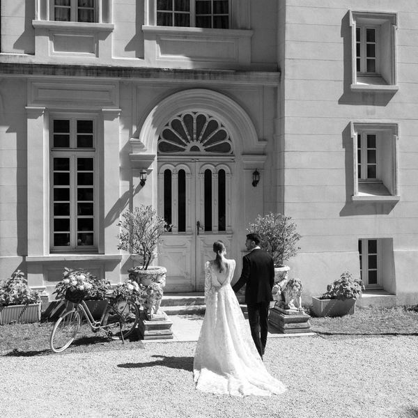 A bride and groom walk hand in hand towards a large, elegant building with arched windows and tall doors. It is a sunny day, and a vintage bicycle and potted plants decorate the entrance. The black and white photo captures a timeless, romantic moment.