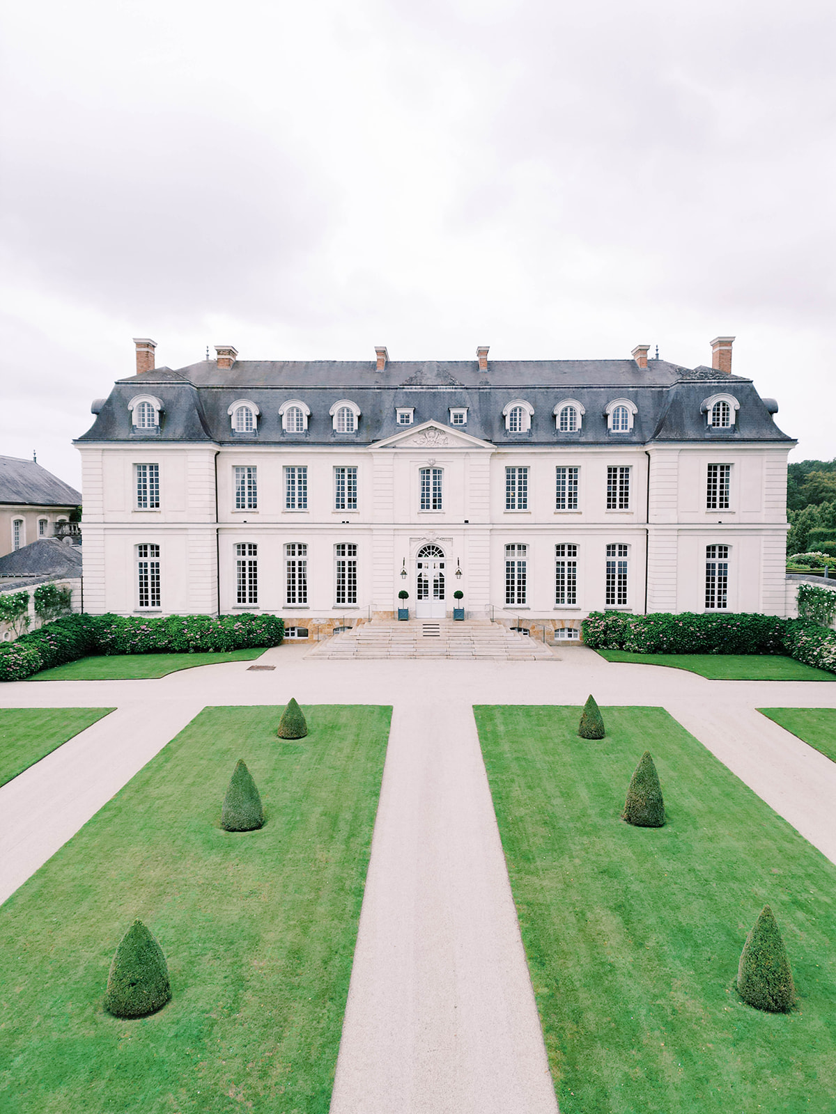 A grand, symmetrical French chateau with multiple windows, a slate roof, and dormer windows sits behind a meticulously manicured garden. The garden features a gravel pathway flanked by neatly trimmed hedges and conical topiaries leading to the chateau’s entrance.