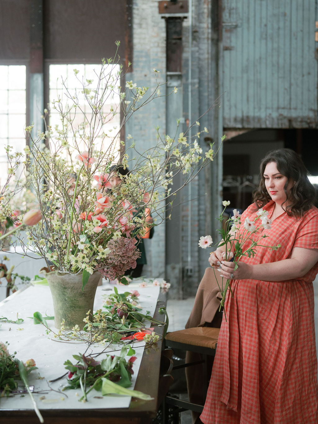 A woman in a checkered orange dress is arranging flowers at a long table inside an industrial space. The table is covered with various flowers and greenery. A large floral arrangement in a vase sits on the table, and sunlight filters through large windows in the background.