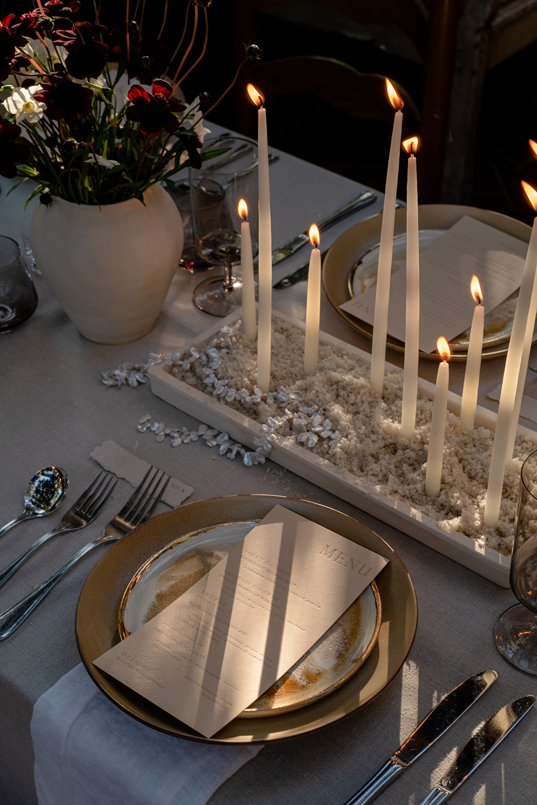 A beautifully set dinner table with glowing tall candles in a rectangular centerpiece filled with white textured material, elegant gold-rimmed plates with menus placed on top, and a vase of dark red flowers. Silver cutlery and glasses are neatly arranged.