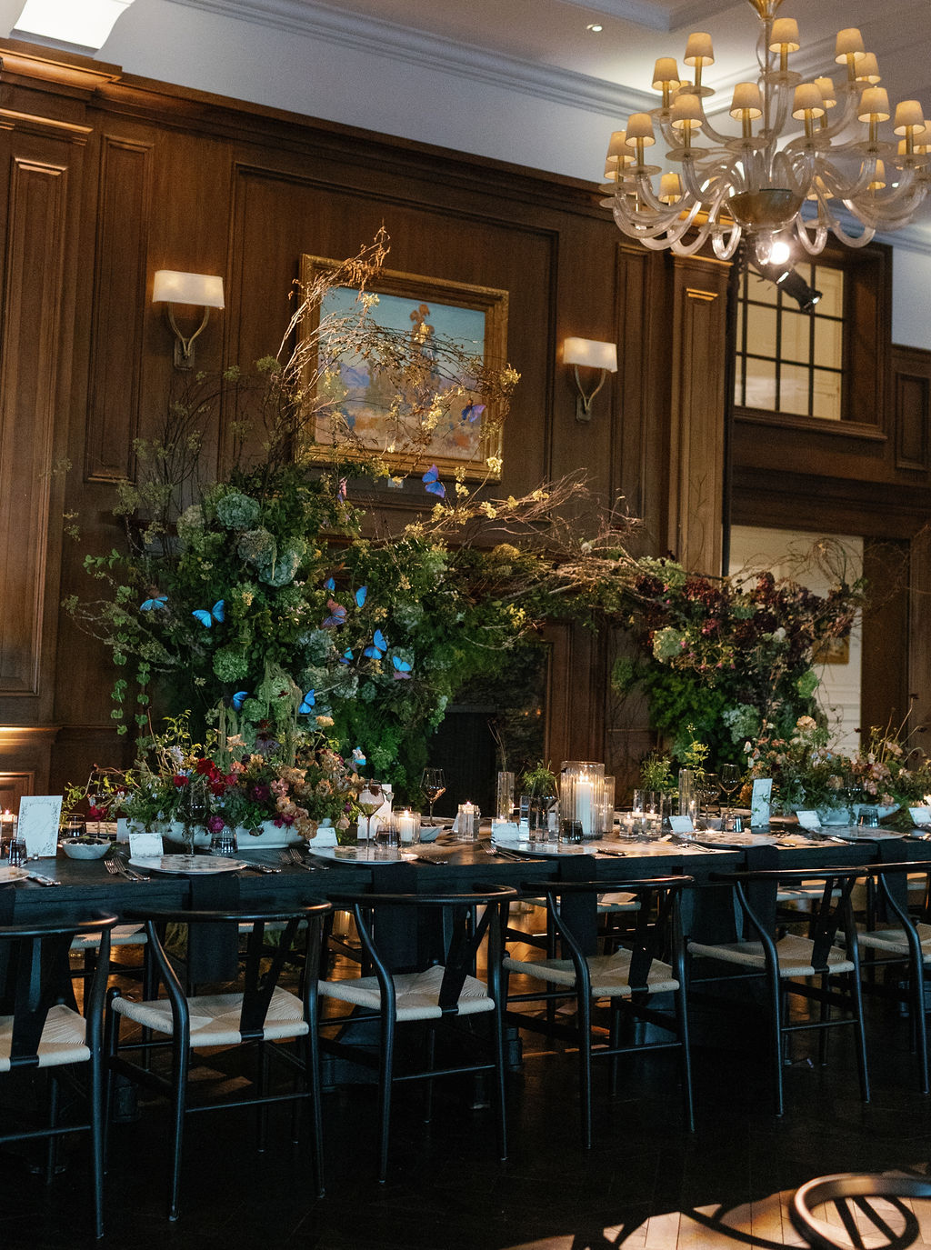 A dining room with a long table set for a formal meal. The table features candles, place settings, and elaborate floral arrangements with greenery and blue butterflies. Wooden paneling, an ornate chandelier, and a painting on the wall enhance the room's elegant atmosphere.