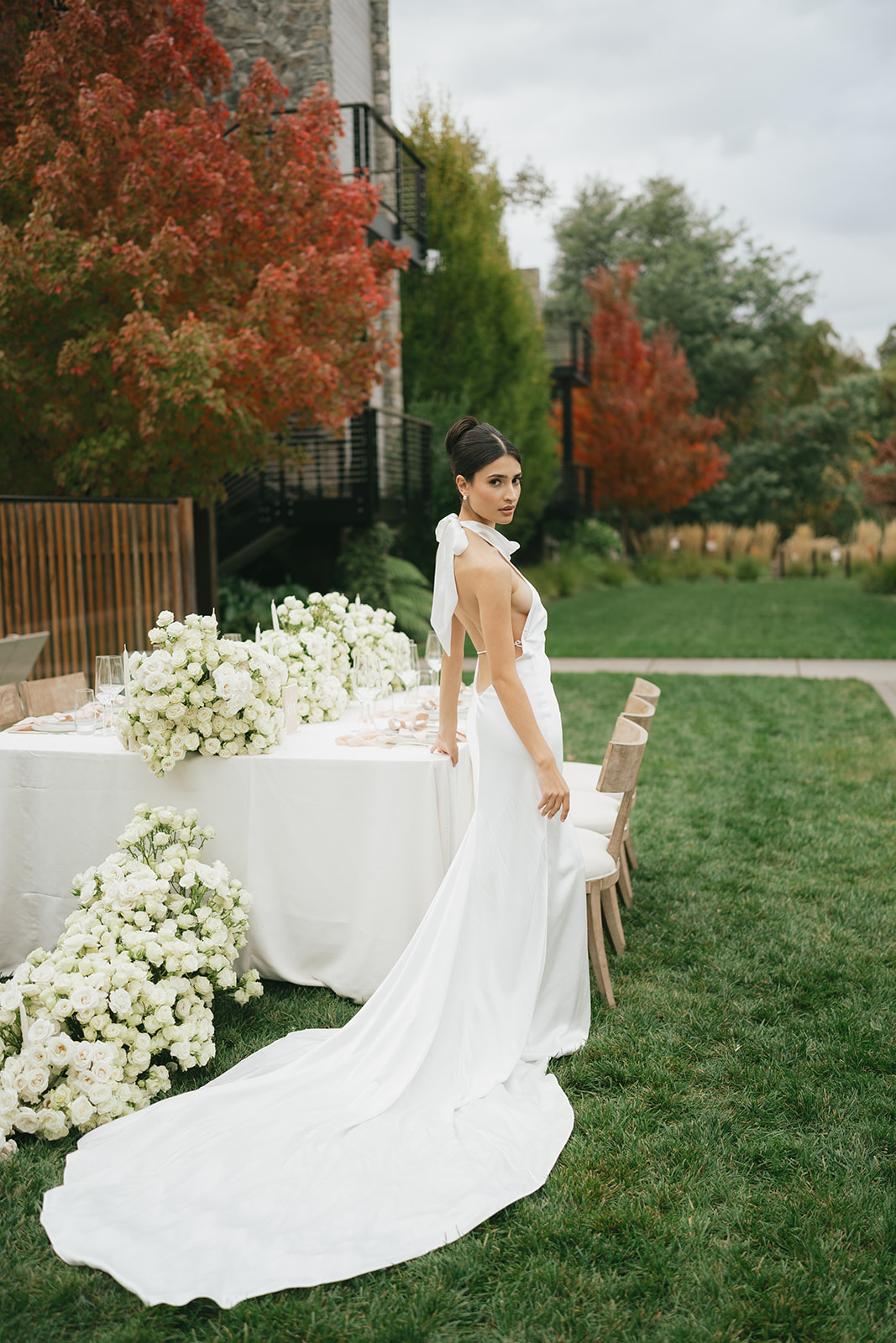 A bride in a white dress stands beside an elegantly decorated outdoor table with white floral arrangements. The table is set on a lush green lawn, with trees showcasing autumn foliage in the background. The bride gazes softly, her dress trailing gracefully behind her.