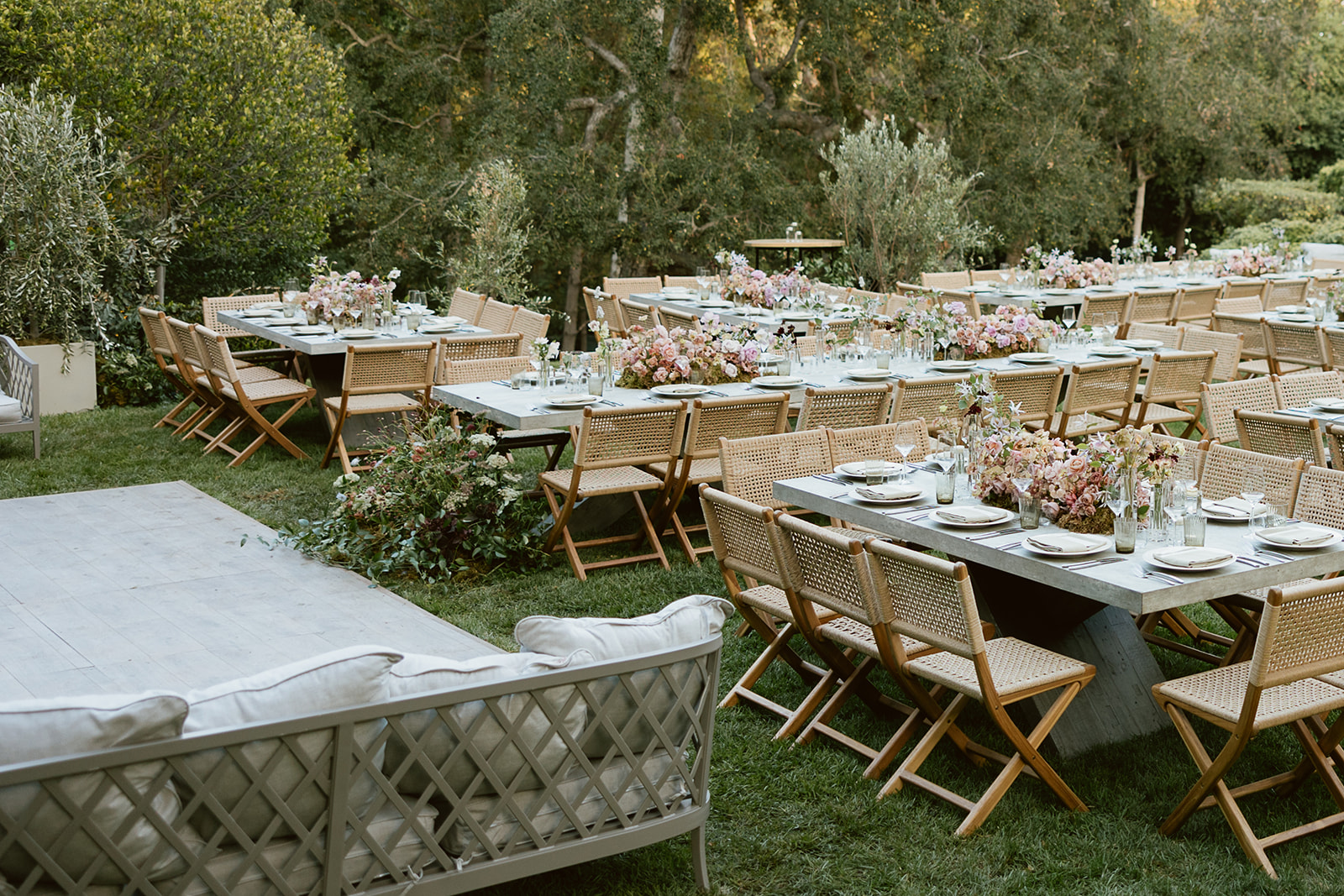 An outdoor event setup featuring long tables with wicker chairs arranged neatly. Each table is adorned with elegant floral centerpieces and set for dinner. The area is surrounded by lush greenery, creating a serene and inviting atmosphere.