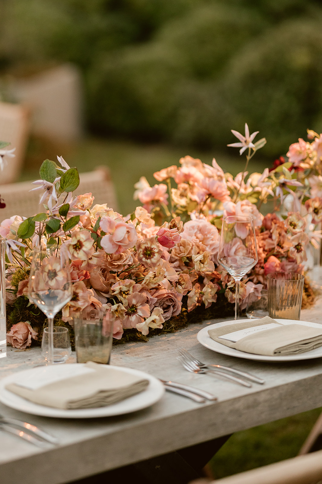 A beautifully set outdoor table adorned with a lush centerpiece of pink and peach flowers. The table features white plates, neatly folded napkins, polished silverware, and elegant glassware, creating a sophisticated and inviting atmosphere.