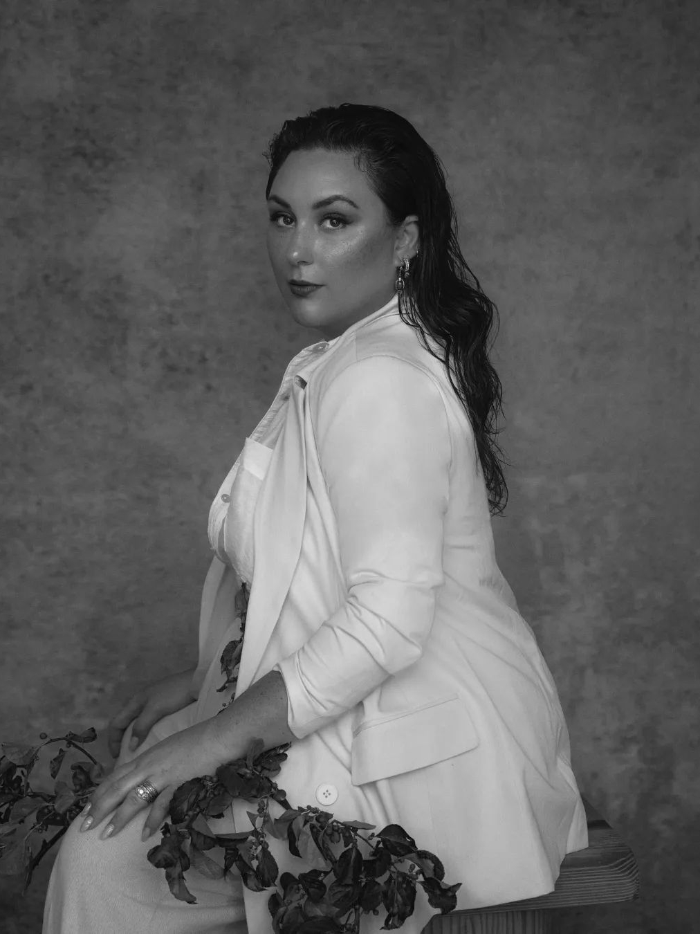 A woman with wet hair, wearing a white blazer over a white shirt, sits on a wooden stool. She faces the camera with a confident expression. Her outfit is accentuated by a leafy branch draped around her. The background is a textured, neutral-toned backdrop.