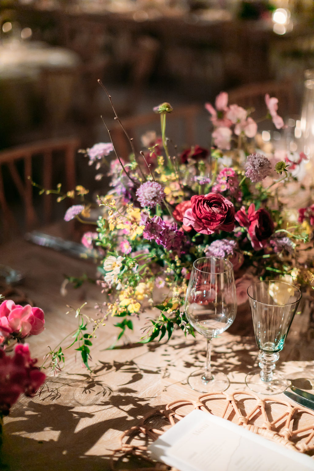 A beautifully set table at a formal event, featuring an elaborate floral centerpiece with red, purple, and yellow flowers. The table includes wine glasses, a folded menu, and elegant cutlery, all softly illuminated by warm, ambient lighting.