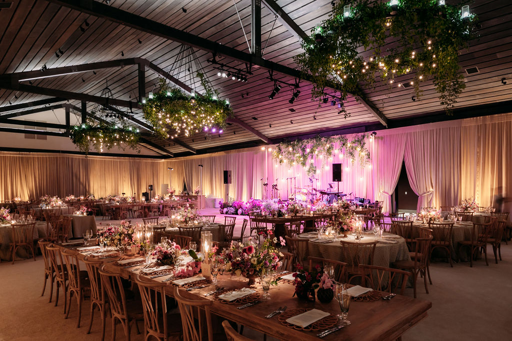 A beautifully decorated event hall set for a formal gathering. Long wooden tables with elegant floral centerpieces, soft pink and purple uplighting, sheer white drapery, and hanging greenery with twinkling lights create a romantic and inviting atmosphere.