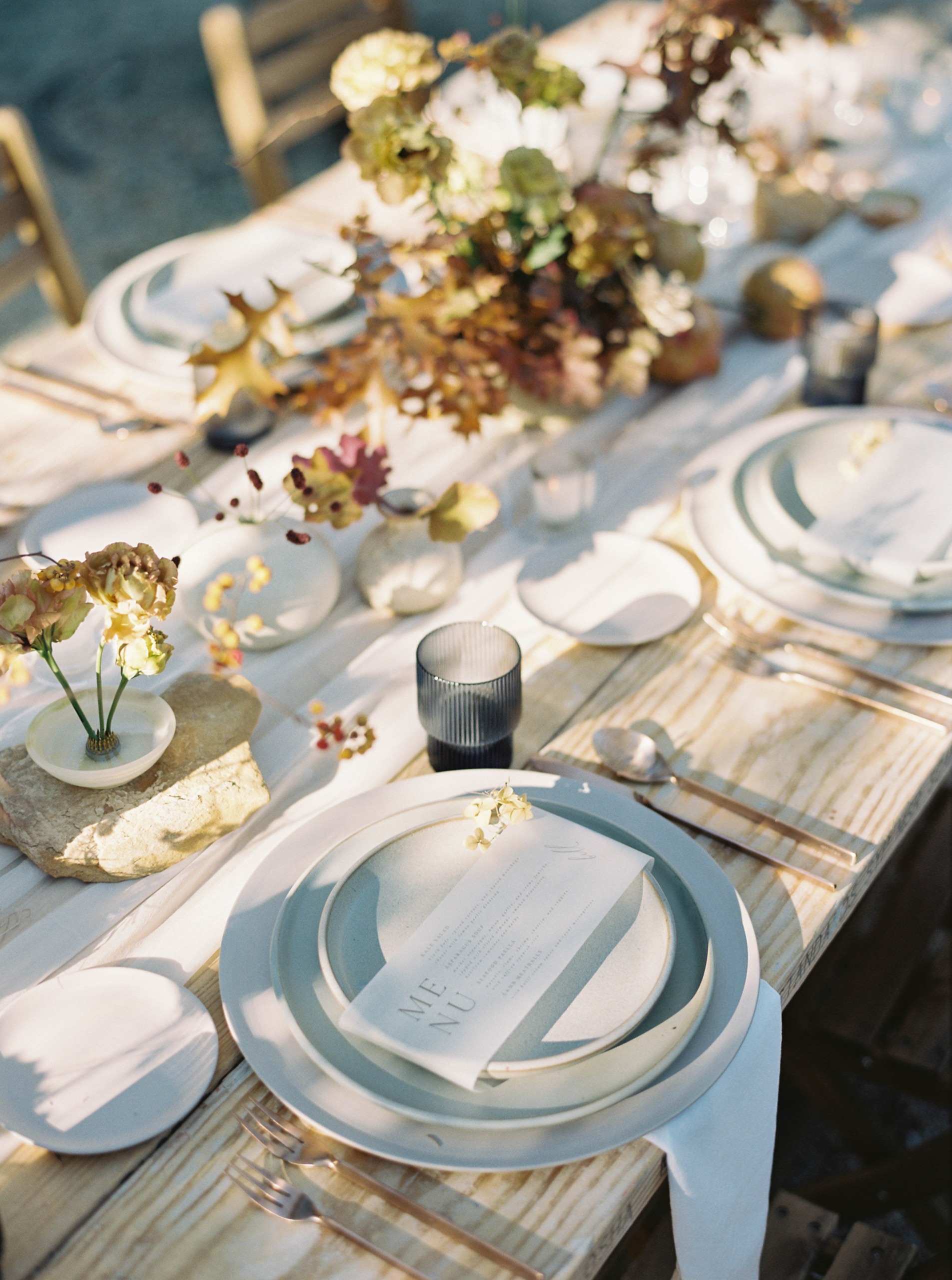 A wooden table is set outdoors with elegant tableware. Each setting includes layered plates, silverware, a menu, and a small flower on the plate. A centerpiece with various flowers and foliage runs the length of the table. Lit candles are placed around.