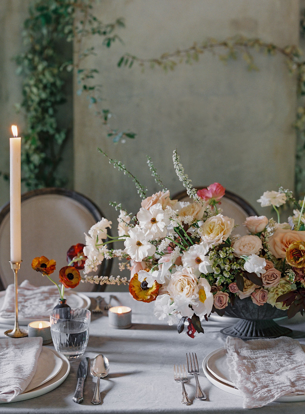 A beautifully set table features elegant floral arrangements with a mix of white, peach, and orange flowers in a gray vase, surrounded by candles. The table is adorned with white plates, silver cutlery, and gray linen napkins, set in a soft, serene ambiance.