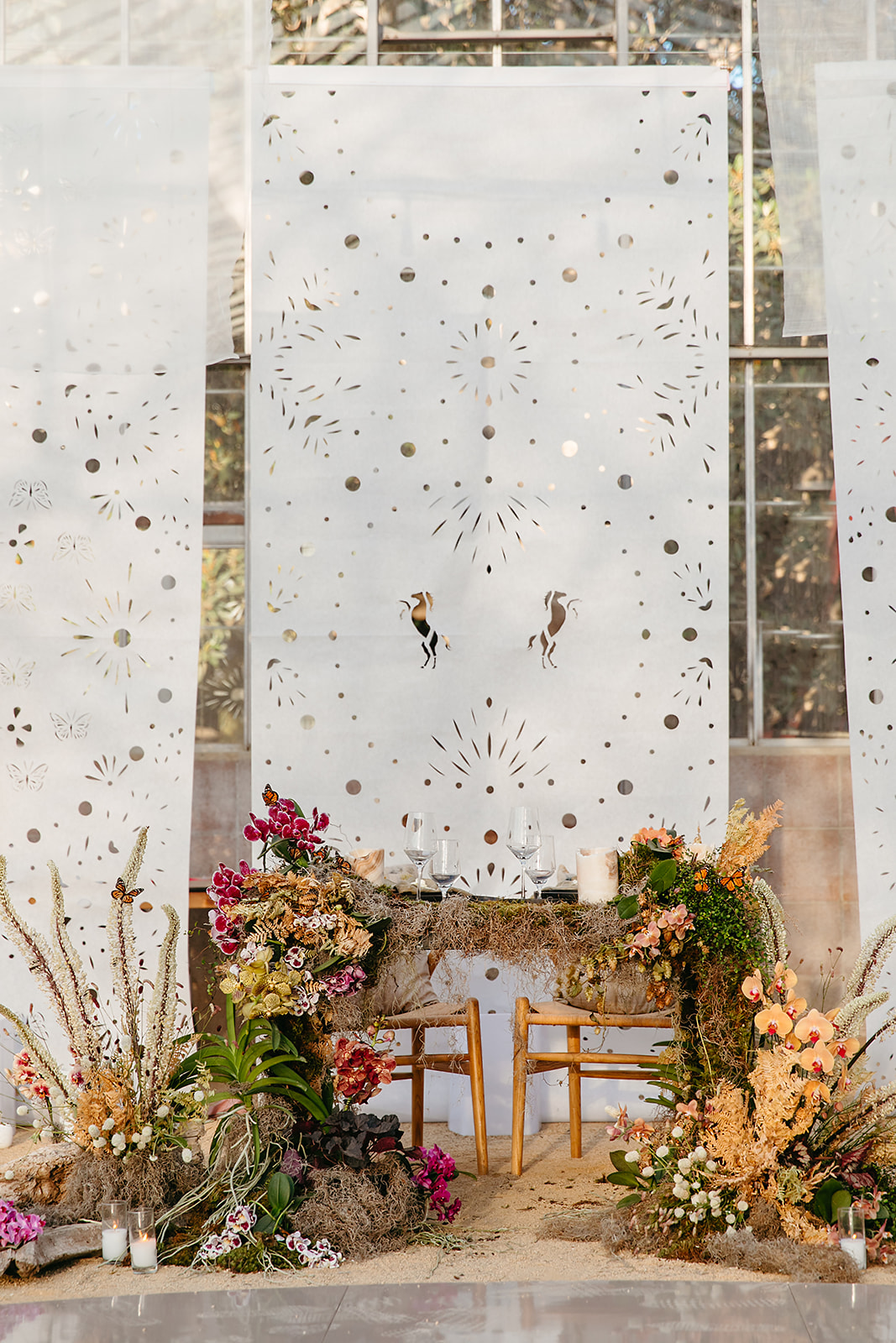 A beautifully decorated wedding ceremony setup featuring two wooden chairs behind a small table adorned with glassware and candles. Surrounding the table are colorful flower arrangements and leafy plants. The backdrop is made of white panels with intricate cut-out designs.