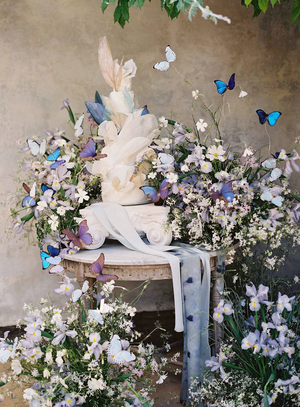 A beautifully decorated table features a white sculptural arrangement surrounded by lush greenery and delicate white and purple flowers. Several blue and white butterflies flutter around, adding a whimsical touch to the serene setting under a leafy canopy.