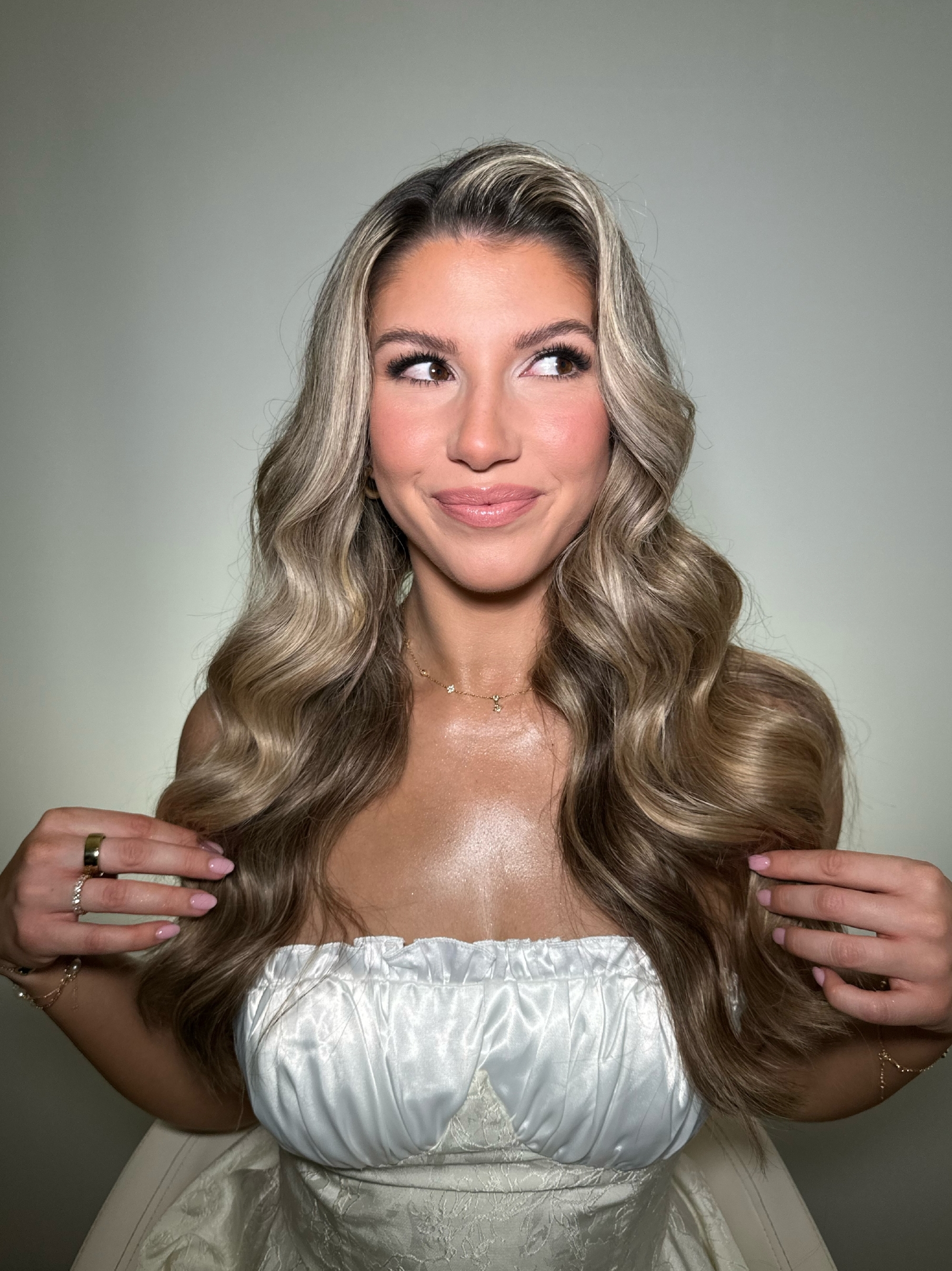 A woman with long, wavy blonde hair is smiling and looking up to her right. She is wearing a white strapless top and has polished nails. She holds a strand of her hair in each hand and has a glowing complexion. The background is plain and light-colored.