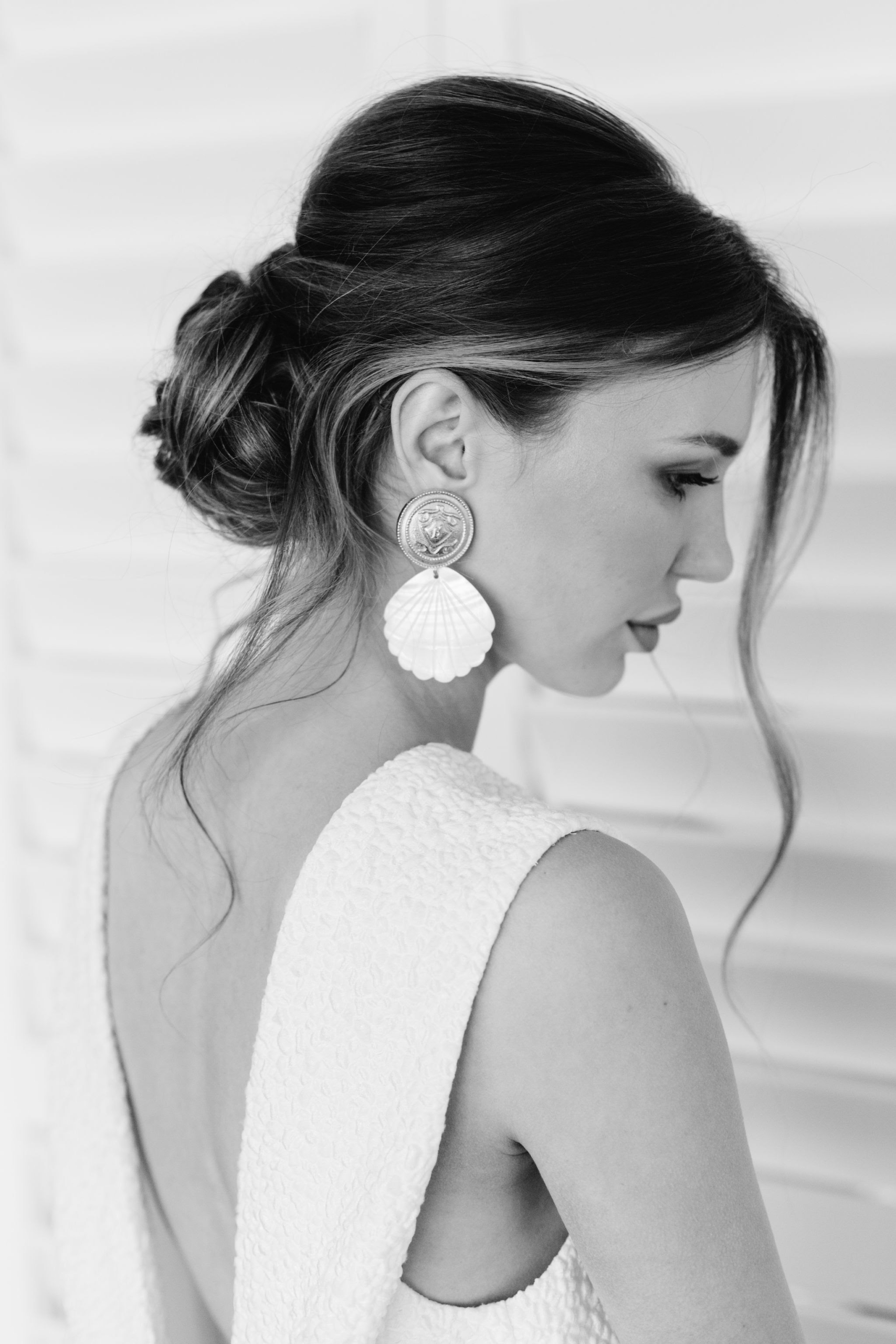 A black and white portrait of a woman with her hair in an elegant updo, featuring a loose strand framing her face. She is wearing a textured, sleeveless dress with a deep back and large, seashell-shaped earrings. She stands against a backdrop of white blinds.