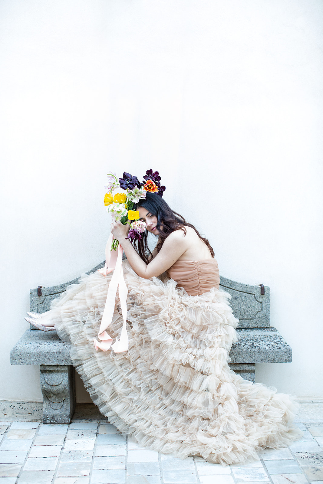 A person wearing a flowing, ruffled gown in a light beige color sits on a stone bench holding a vibrant bouquet of flowers. The background is a plain white wall and the person appears to be smiling, with their head slightly tilted to one side.