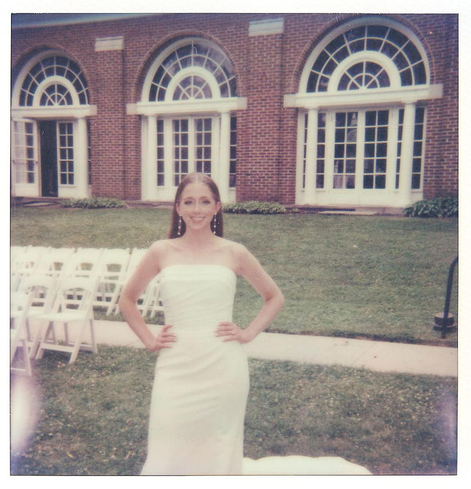 A woman in a strapless white dress stands with her hands on her hips in front of an elegant building with large arched windows. She is smiling and posing for the camera on a grassy area with white folding chairs arranged nearby.