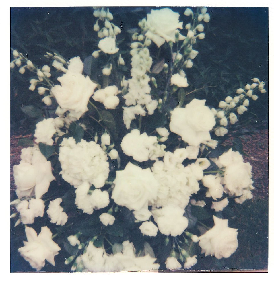 A close-up Polaroid photograph of a floral arrangement featuring various white flowers, including roses and peonies, with green foliage in the background. The flowers are arranged in a symmetrical and lush display.