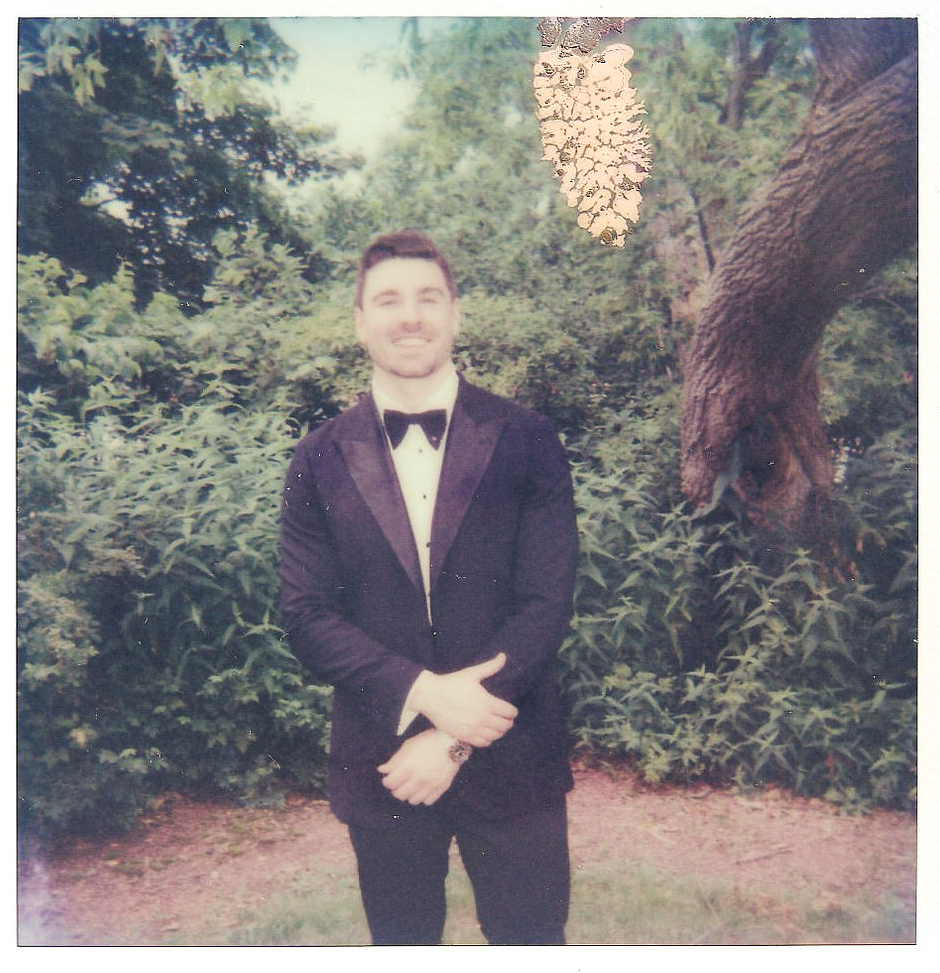 A man stands outdoors in front of green foliage, wearing a black tuxedo with a white shirt and black bow tie. He has short hair and a beard, and he smiles while holding his hands clasped in front. A portion of the image is damaged at the top center.