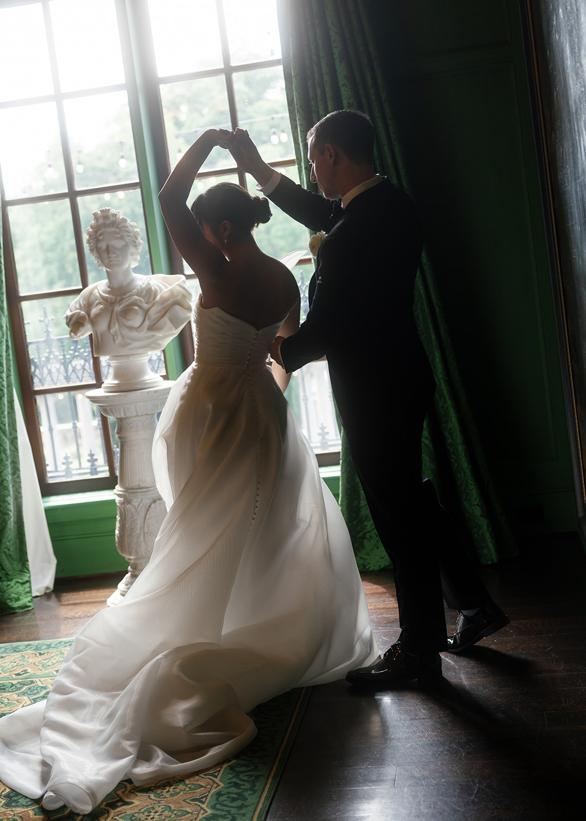 A bride and groom dance near a large window in a dimly lit room. The bride, wearing a white gown, is gracefully twirling as the groom in a dark suit holds her hand. A marble statue is visible in the background by the window. Green drapes frame the scene.