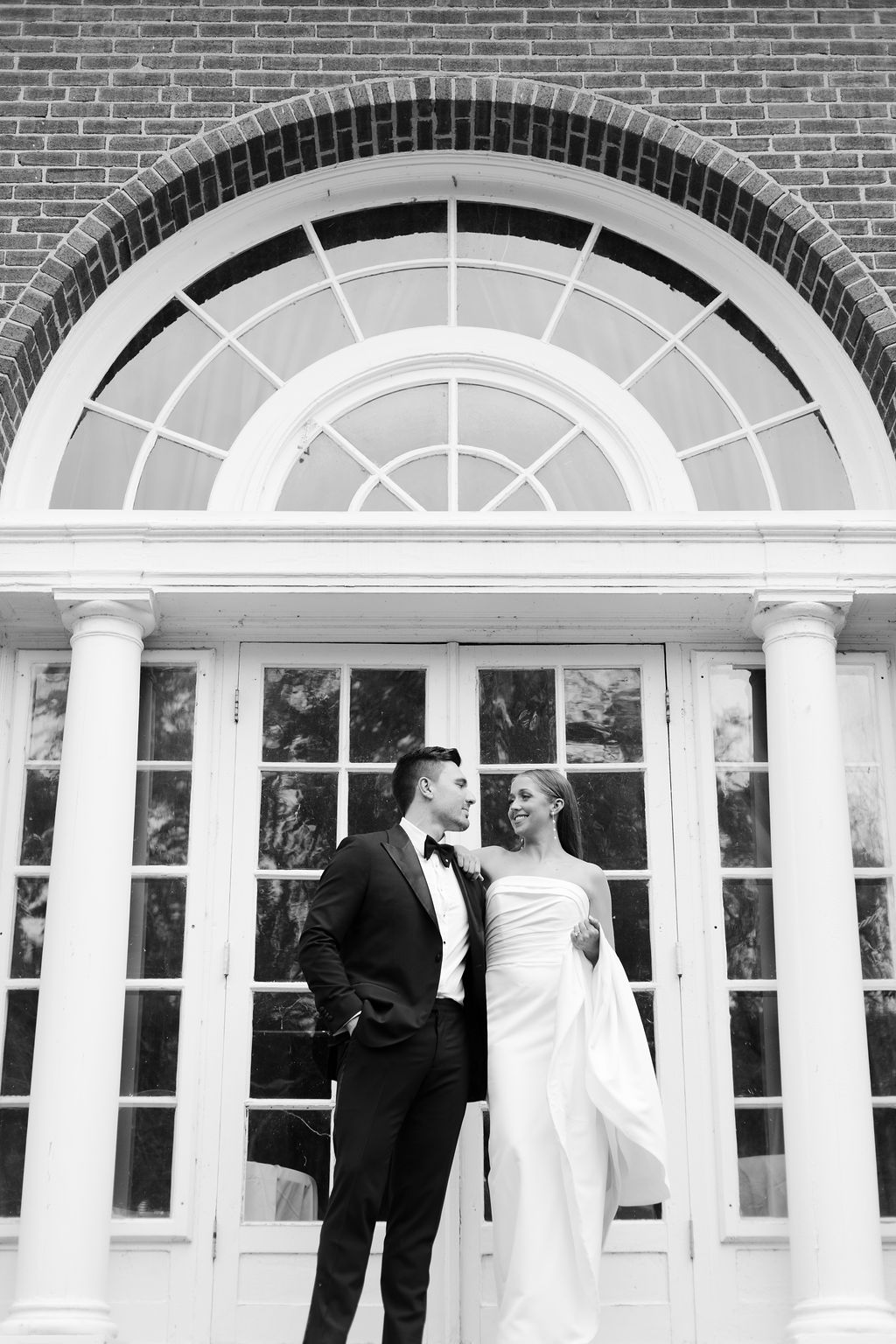 A bride and groom stand together in front of a large, arched window with a brick surround. The groom is wearing a black tuxedo, and the bride is in a white strapless gown, holding the train. They look at each other affectionately. The photo is in black and white.