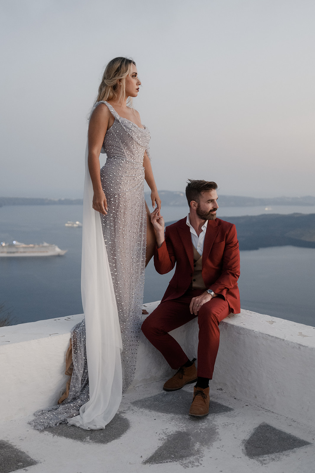 A woman in a silver, sequin evening gown stands next to a man sitting on a white wall, with the man holding her hand. He is dressed in a red suit. They are overlooking a body of water with hills in the background, under a dusky sky. A cruise ship is visible in the water.