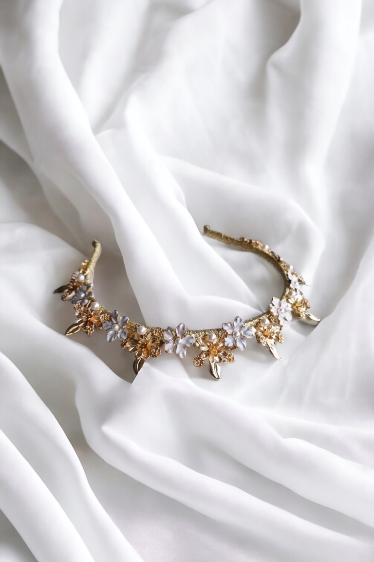 A delicate, gold-colored headband adorned with small, intricate floral designs in silver and champagne hues is elegantly laid on soft, white, draped fabric.