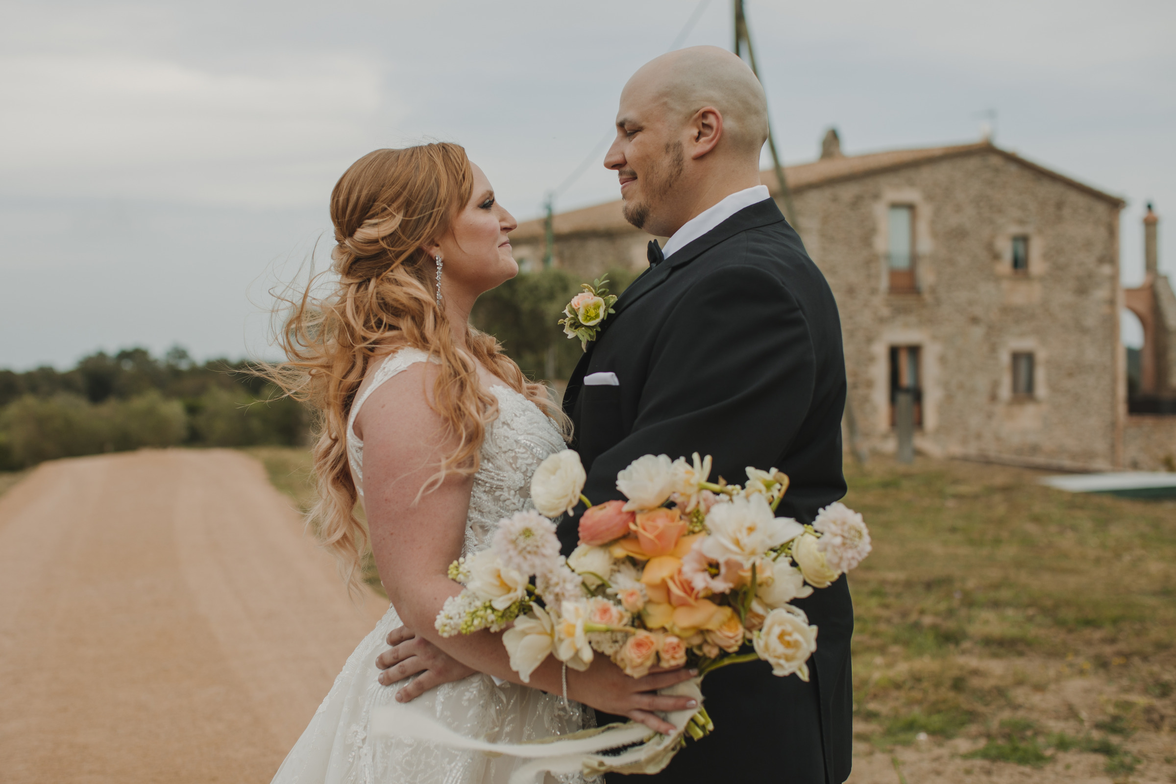 A bride with long red hair in a white lace wedding dress and a groom with a bald head in a black suit stand facing each other, smiling. The bride is holding a colorful bouquet of flowers. A rustic stone building and a dirt path are visible in the background.