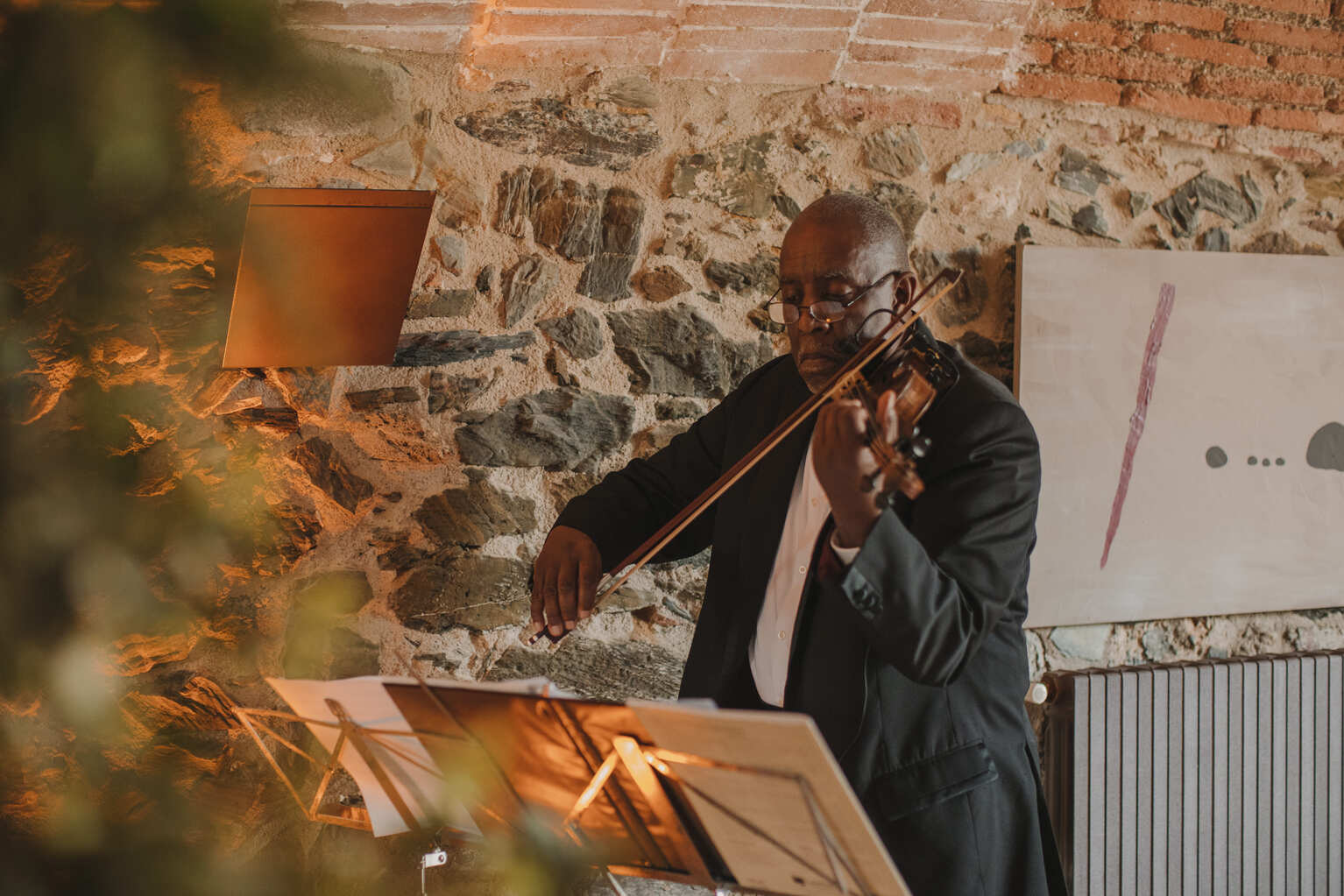 A man in a black suit and glasses plays a violin in an indoor setting with stone walls and modern light fixtures. Sheet music is on a stand in front of him. The background includes abstract art and a dimly lit sconce.