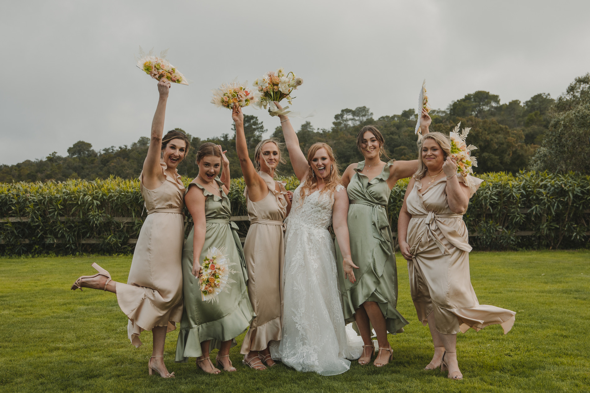 A bride in a white gown stands joyfully with five bridesmaids wearing green and beige dresses. They are outdoors on a lush lawn with trees in the background, holding bouquets aloft and smiling happily. One bridesmaid is playfully lifting her leg.