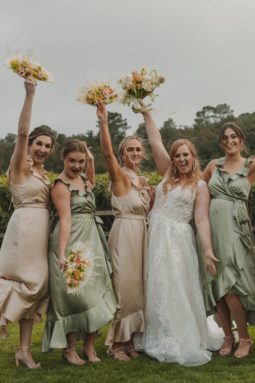 A bride in a white lace dress poses joyfully with four bridesmaids in beige and green dresses. Each bridesmaid holds a bouquet and raises an arm in celebration. The backdrop features lush greenery and trees, adding to the festive outdoor setting.