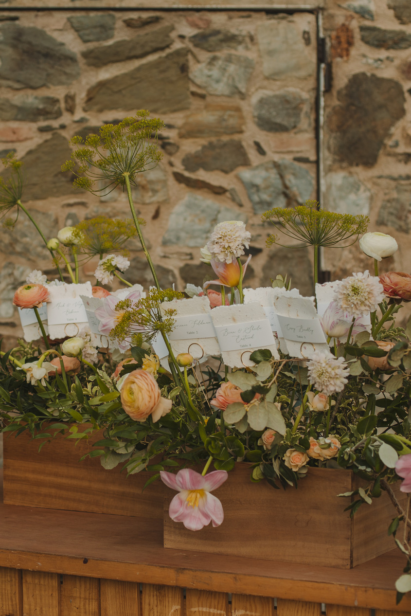 A rustic display of three wooden boxes filled with various pastel-colored flowers, including roses and peonies, some with handwritten notes attached. The backdrop is a textured stone wall, and there is greenery arching from both sides of the image.