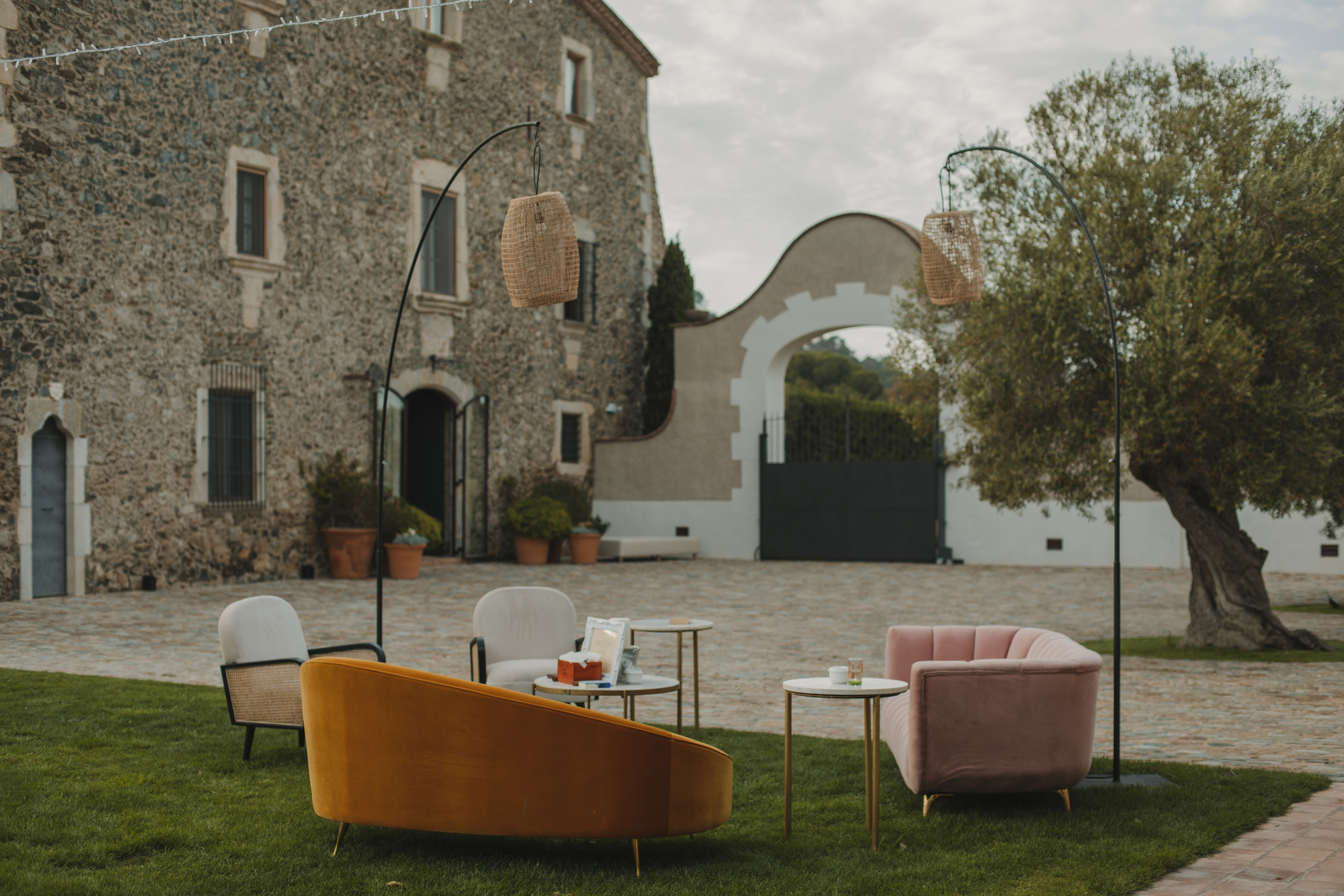 An outdoor seating area with a mustard yellow sofa, pink armchair, gray chairs, and round tables on a grassy lawn. Two tall lamps with woven shades stand next to the seating. A rustic stone building and arched gate are in the background.