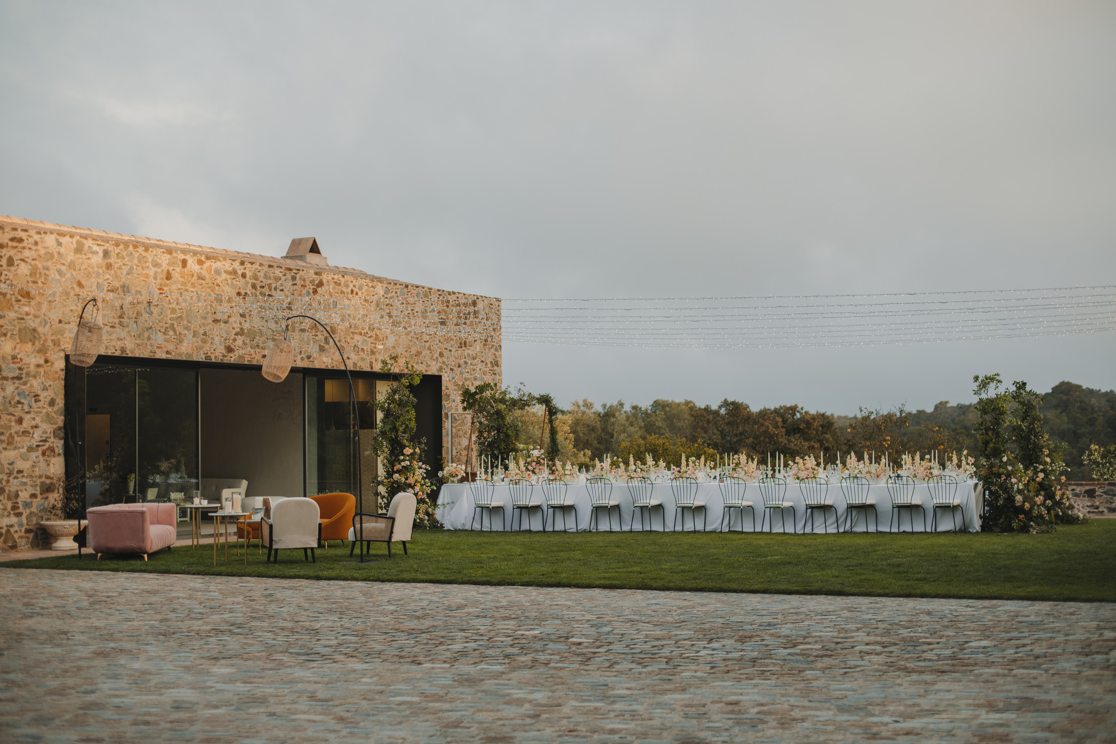 An outdoor dining setup beside a rustic stone building. A long table with white tablecloths, elegant table settings, and floral centerpieces is arranged on a green lawn. Cozy seating area with colorful chairs is adjacent. String lights are hung above.