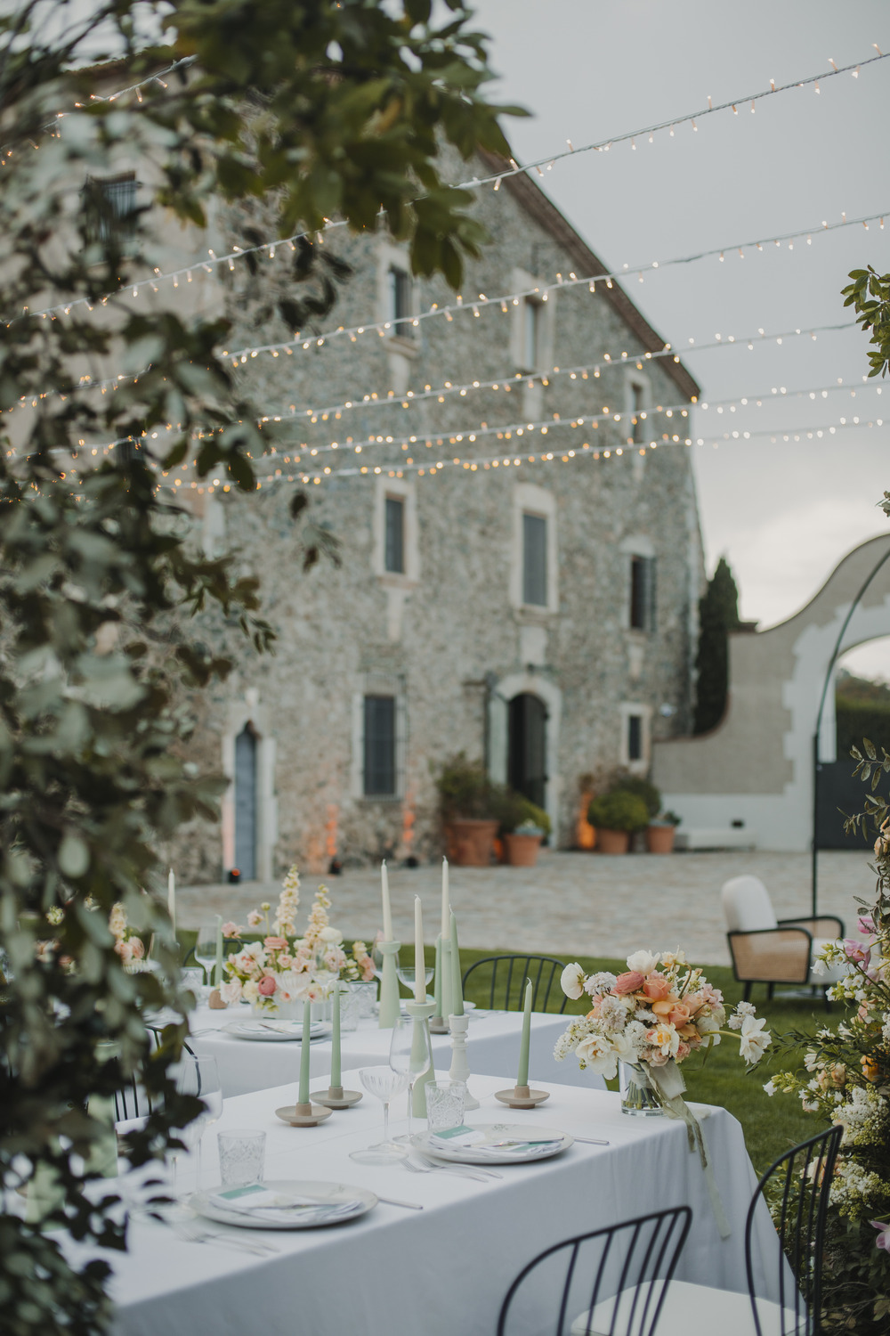 A beautifully set outdoor dining area with a white tablecloth and elegant floral arrangements. The scene is framed by a rustic stone building, and string lights hang overhead, adding a warm ambiance to the setting. Green candles and neatly set plates complete the table.