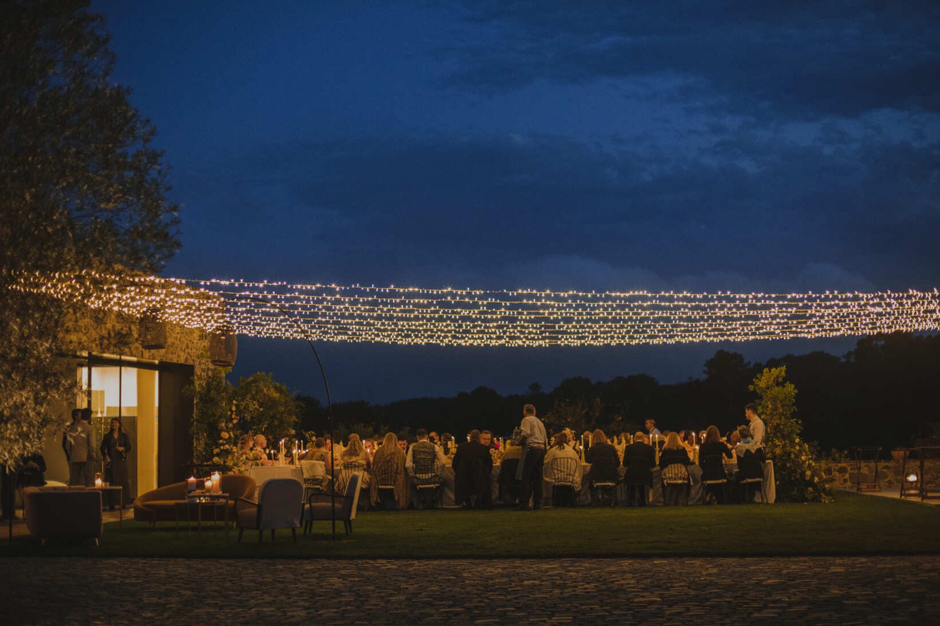 A group of people are gathered outdoors under string lights in the evening. The scene includes tables and chairs set up for a likely event or dinner, with a dimly lit building to the left and surrounding greenery creating a serene atmosphere.