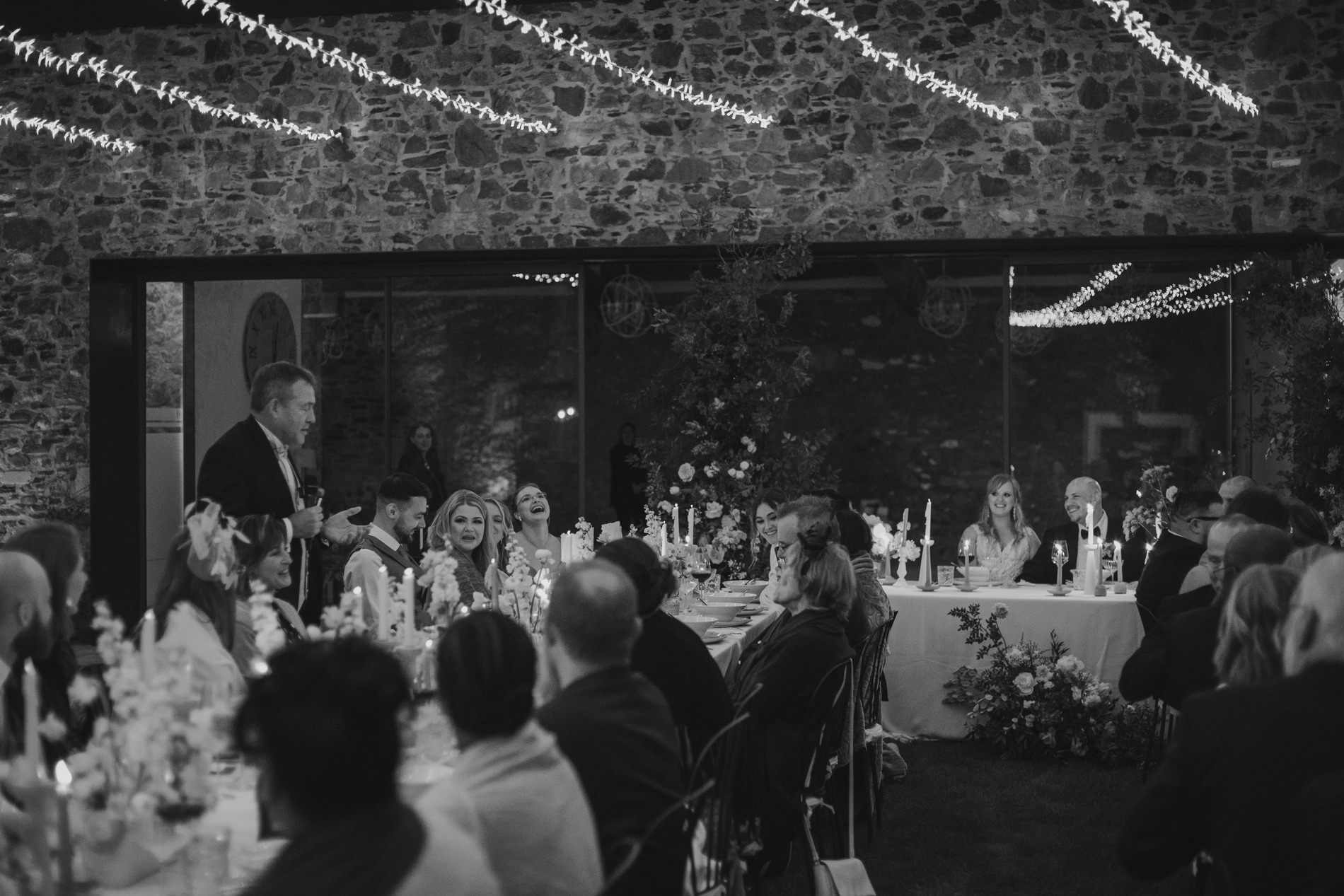 A black and white photo of a wedding reception. Guests are seated at long tables adorned with floral arrangements and candles. A man stands giving a speech, while others, including the bride and groom, listen. String lights hang from the ceiling, illuminating the stone walls.