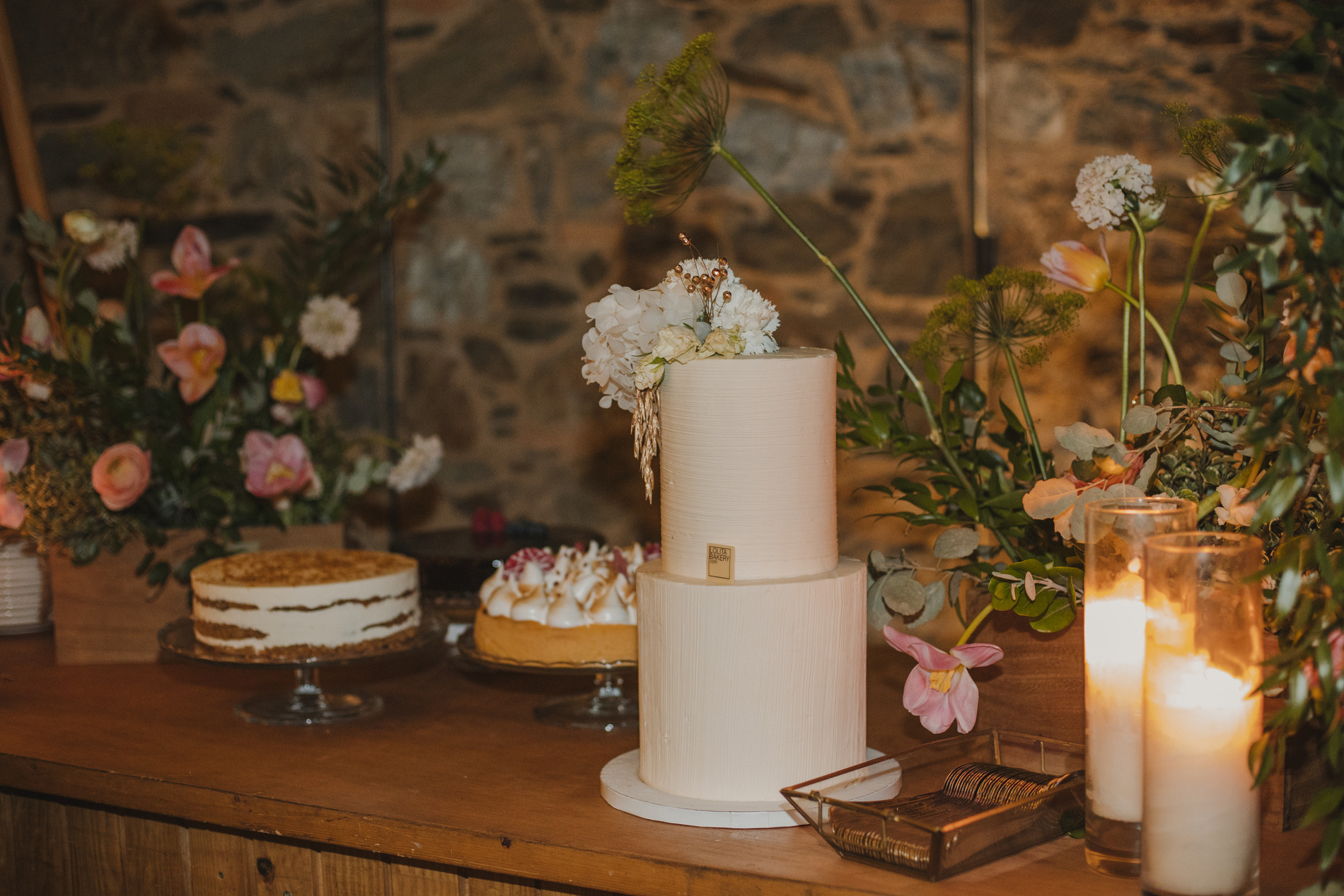 A table is adorned with a two-tiered white wedding cake topped with flowers, placed against a rustic stone wall. Surrounding the cake are various other cakes and desserts, floral arrangements, and lit candles, creating a charming and cozy ambiance.