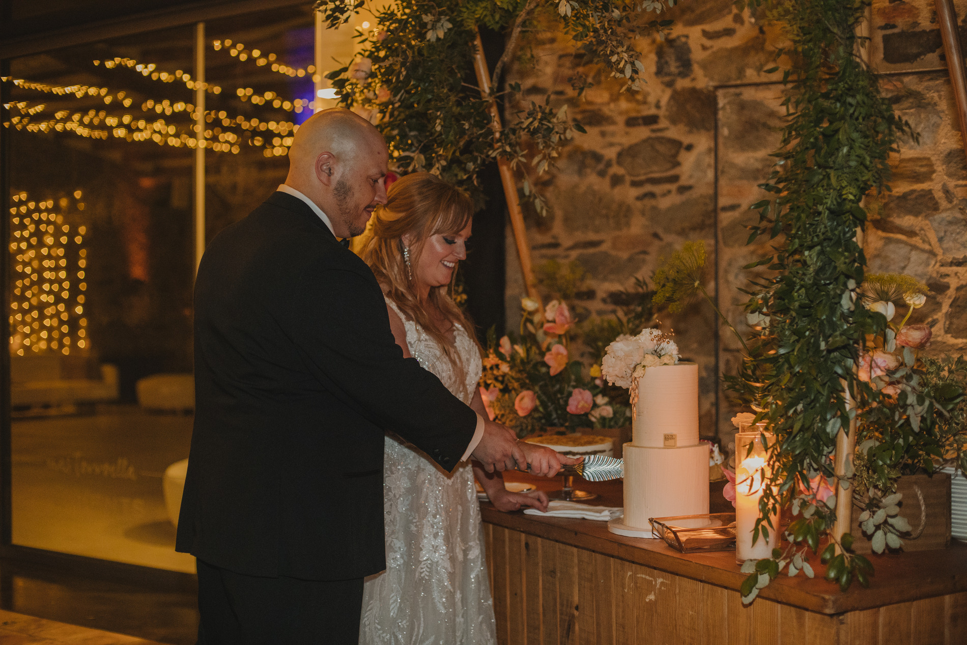 A bride and groom stand together in front of a rustic wooden table, smiling as they cut into a white wedding cake adorned with floral decorations. Behind them, a stone wall is partially covered with greenery and flowers, and fairy lights twinkle in the background.