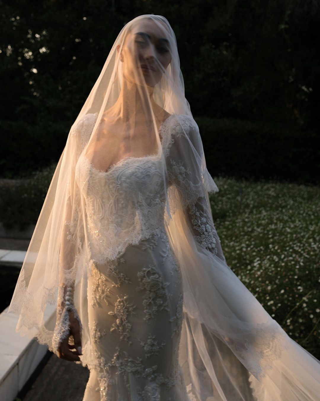 A bride stands in an elegant lace wedding dress, partially illuminated by soft light. She holds out the sheer veil that covers her, creating a flowing and ethereal effect. A green, floral backdrop adds a natural element to the scene.