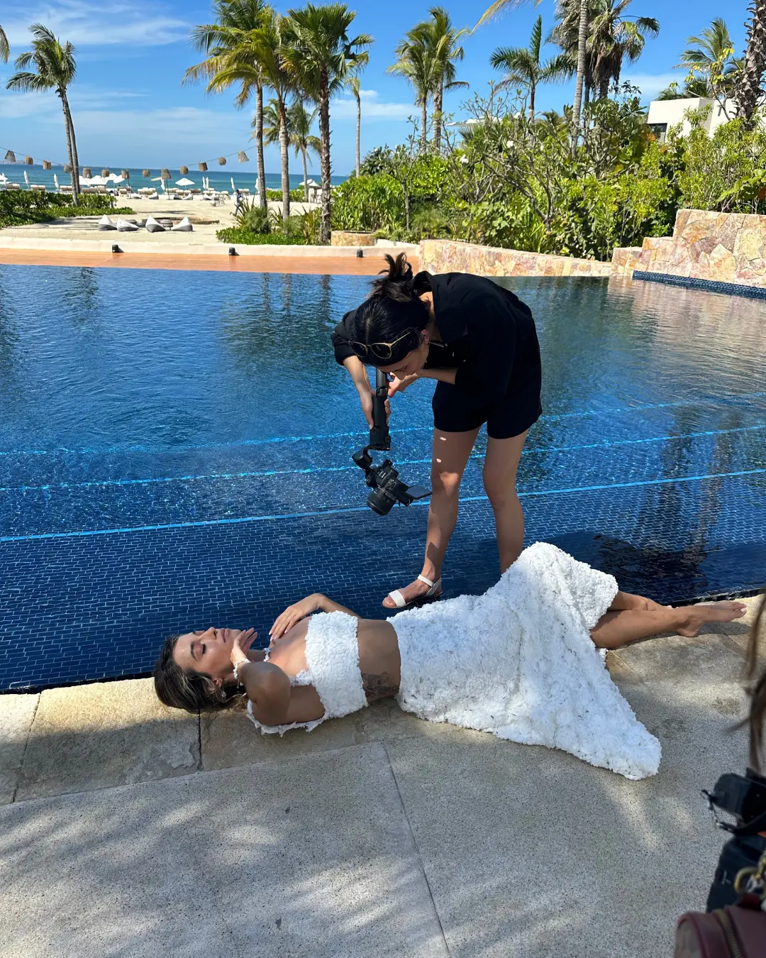 A woman in a white outfit lies on her back near a pool, posing for a photographer. The photographer, dressed in black, crouches down to take her photo. Palm trees and beach chairs are visible in the background, hinting at a tropical setting.